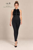 A woman modeling a sleeveless black top and slim black pants against a plain beige studio background. Text in the upper left reads 'UK 8 Apple shape Slightly mid section fullness'.